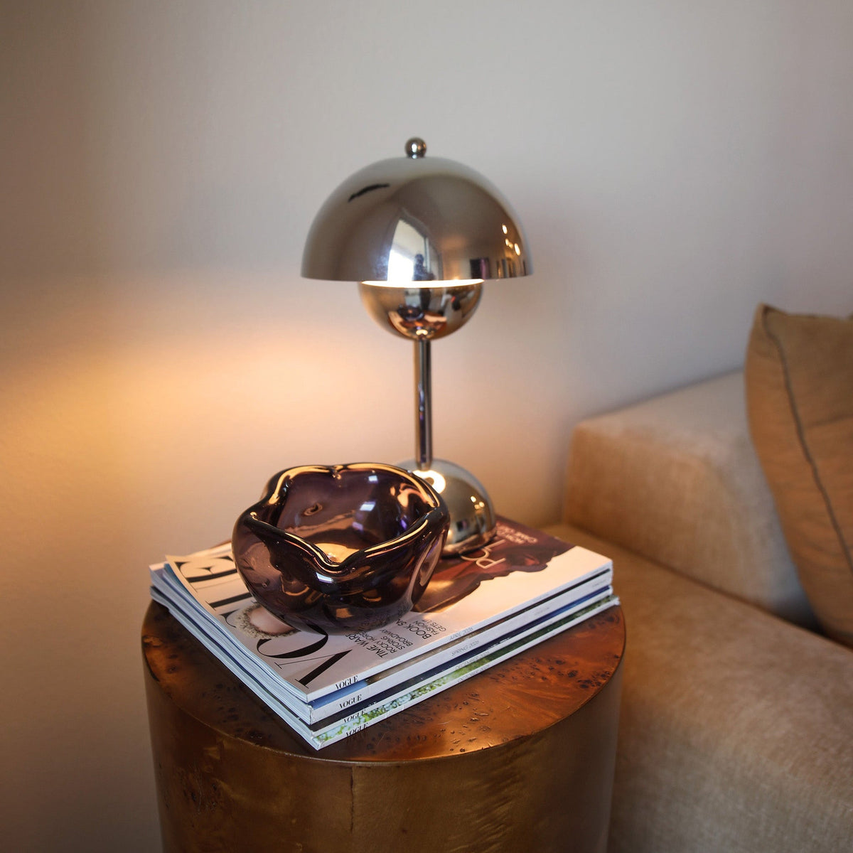 A polished silver dome lamp sits on top of a stack of magazines on a round wooden side table, next to a beige sofa in a dimly lit room.