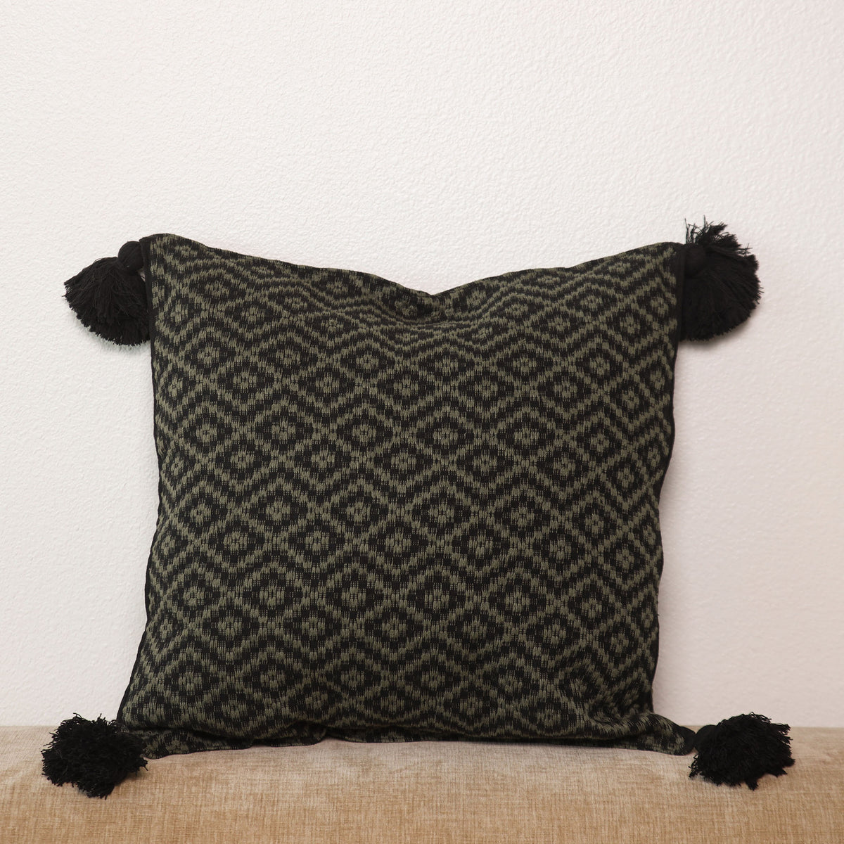 A square throw pillow with a black and dark gray geometric woven pattern and black pom-pom tassels on all four corners, resting on a beige textured surface against a white wall.