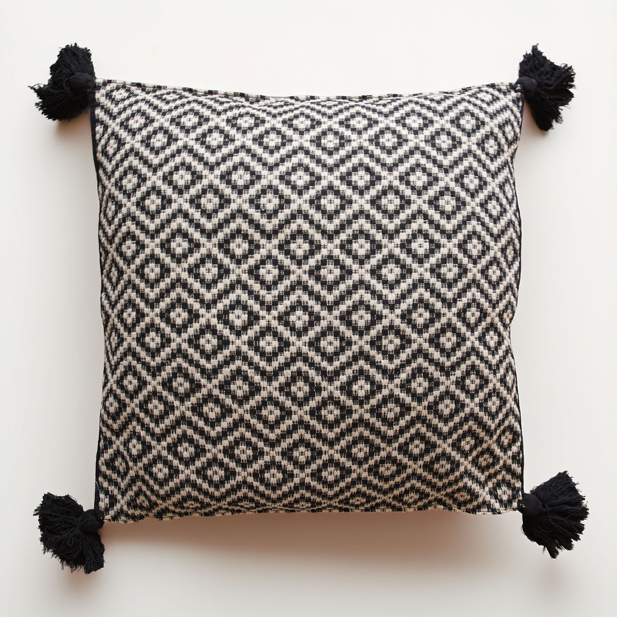 A square throw pillow featuring a black and off-white woven geometric diamond pattern, finished with four black tassels at the corners, shown against a plain white background.