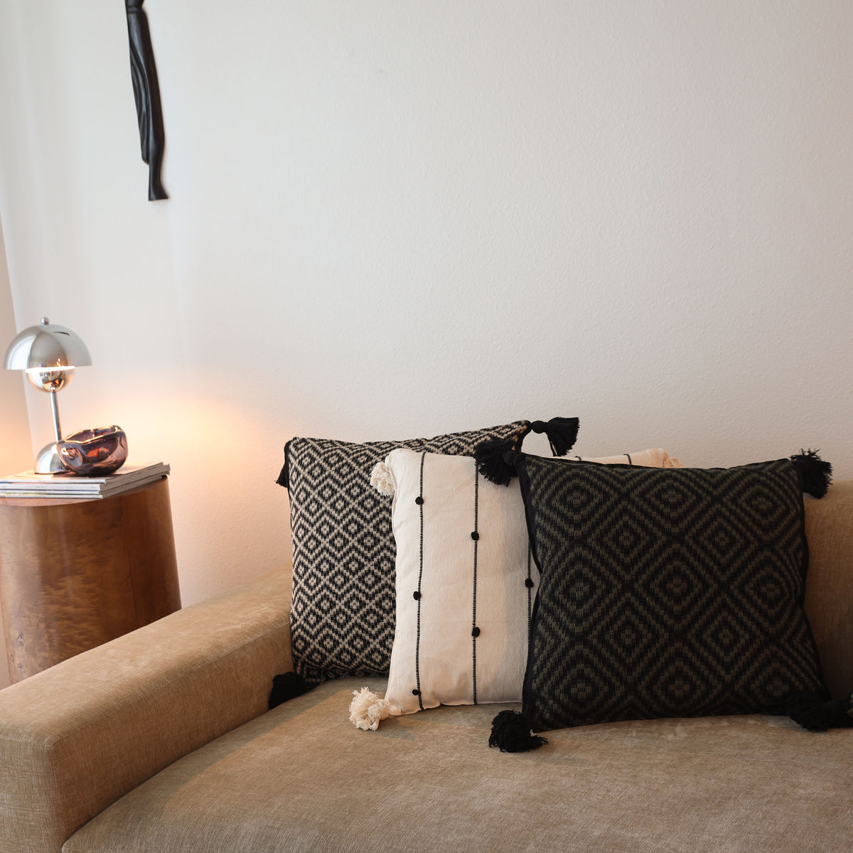 Three decorative throw pillows styled on a beige fabric sofa in a living room setting. The pillows include a black and white geometric patterned pillow, a cream and white vertical striped pillow with black tassels, and a black pillow with a geometric pattern and black pom-poms.
