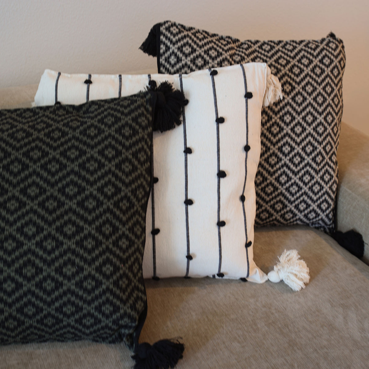 Three decorative throw pillows arranged on a beige sofa: one black-and-white geometric pattern pillow with black tassels, one white pillow with vertical black stripes and black pom-poms plus a white tassel, and one black-and-white geometric pattern pillow with black tassels.