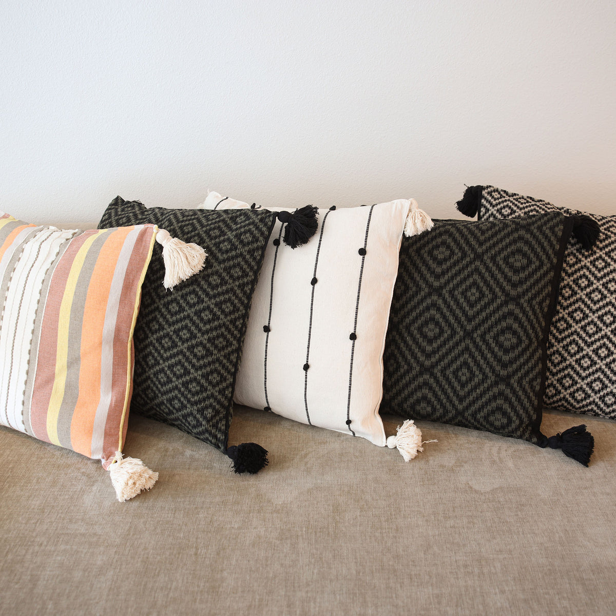 A set of decorative throw pillows arranged on a beige fabric sofa against a plain white wall. The pillows include a striped pillow with cream, orange, and gray tones, a black geometric pattern pillow, a white pillow with black vertical lines and tassels, and other black patterned pillows.