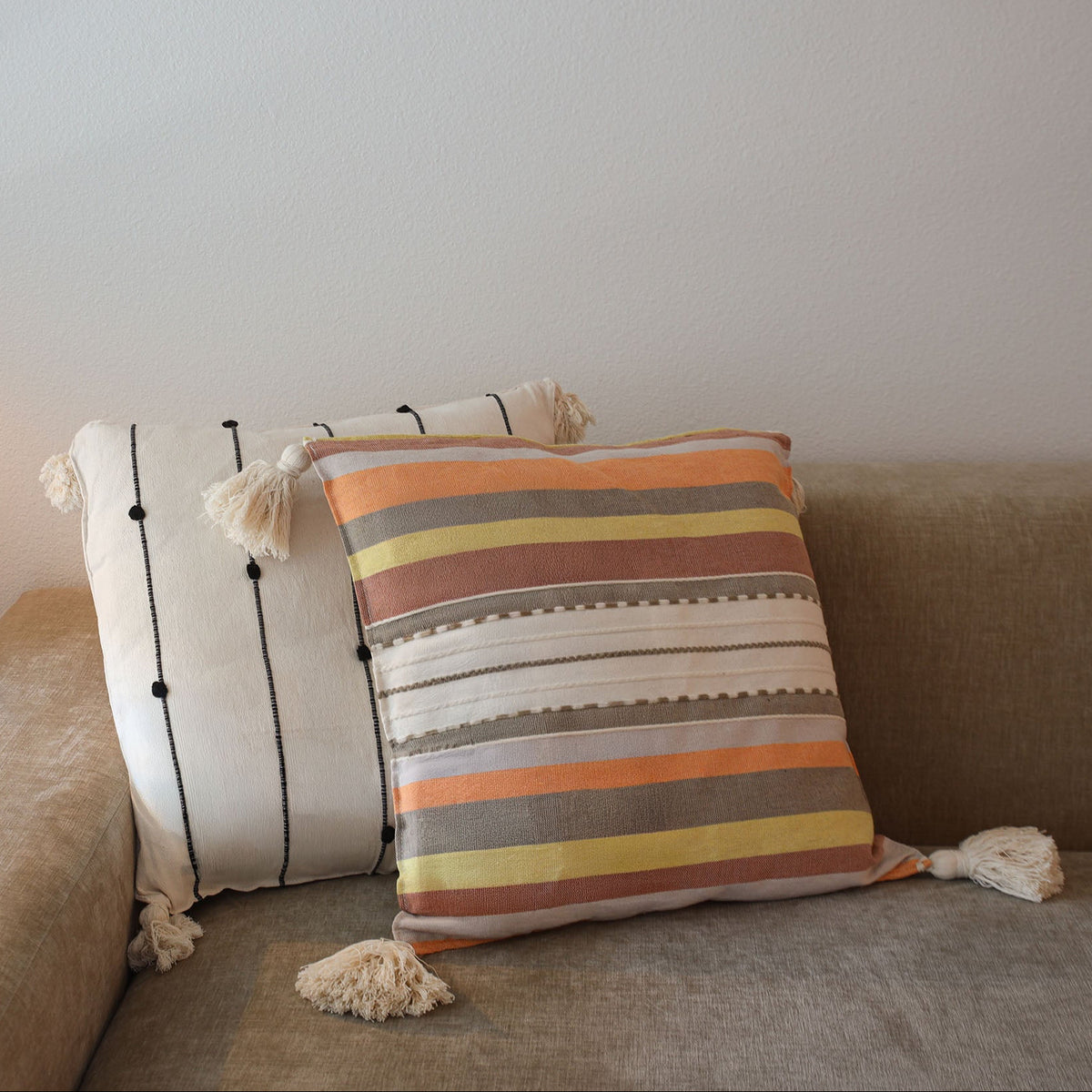 Two decorative throw pillows on a beige fabric sofa against a plain light wall. The front pillow has horizontal stripes in orange, yellow, brown, cream, and gray with tassels, and the back pillow has vertical black stripes on a cream background with tassels.