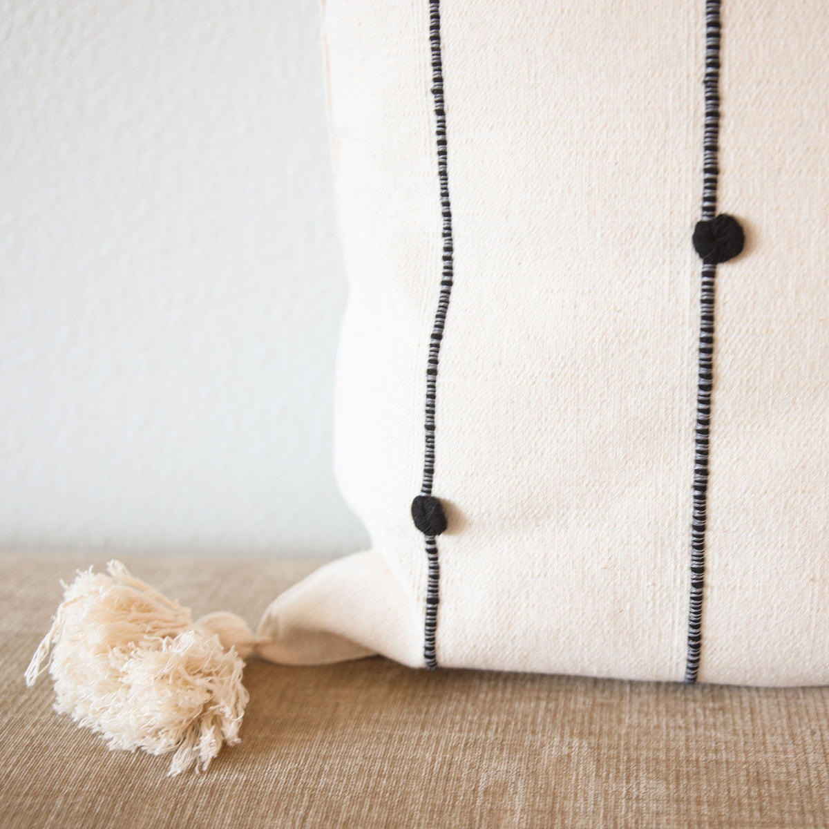 A close-up of a cream-colored woven throw pillow with black embroidered vertical lines and black pom-poms, featuring a matching cream tassel, resting on a beige textured surface against a light gray wall.