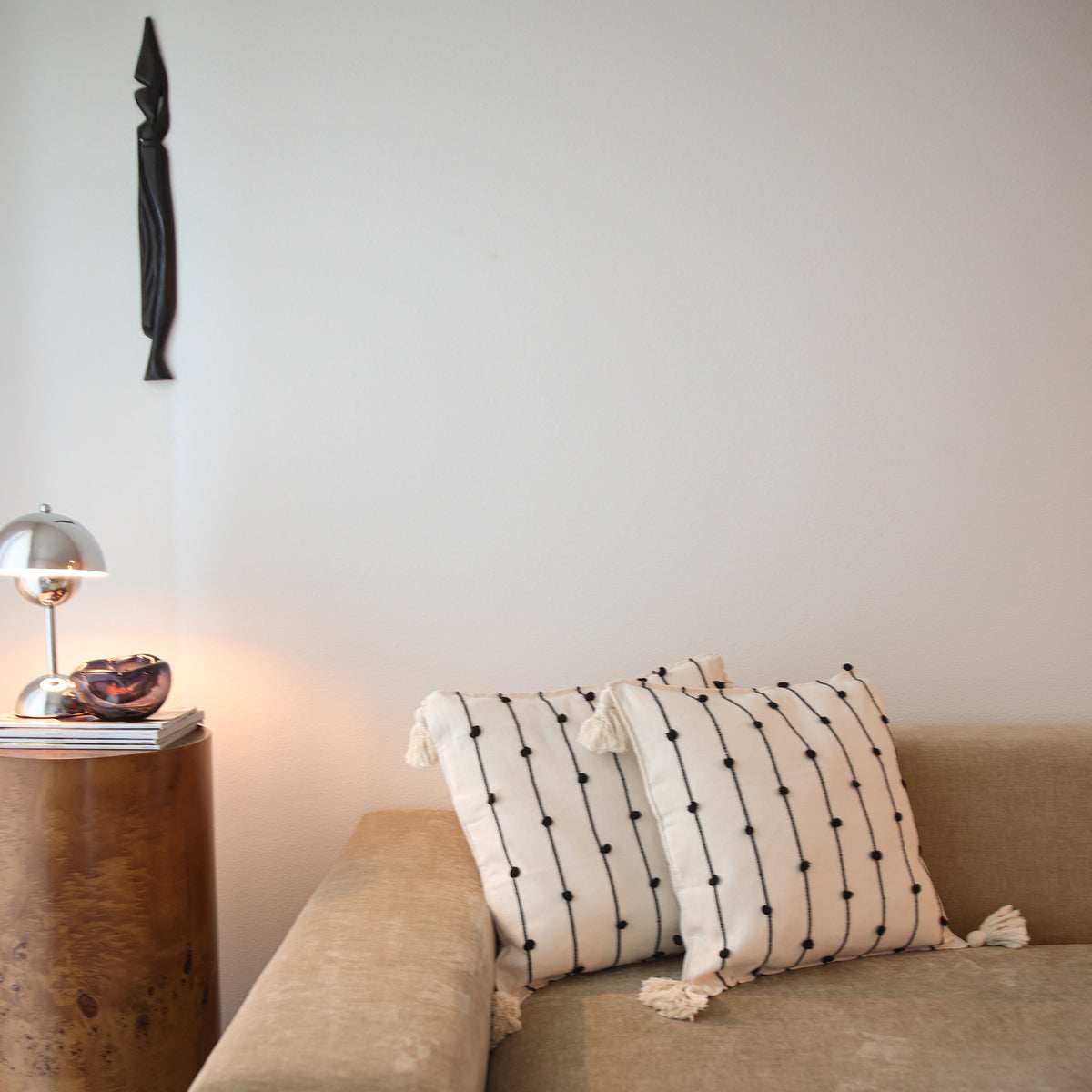 Two cream throw pillows with black embroidered vertical lines and tassels resting on a beige sofa next to a wooden side table with a silver lamp.