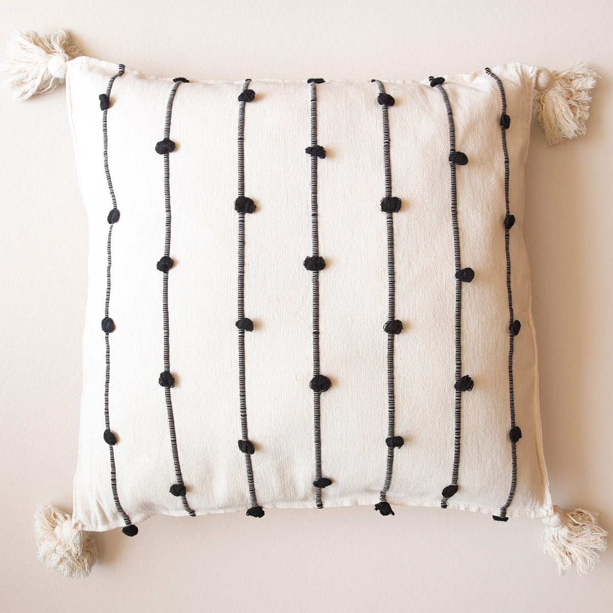 An off-white textured throw pillow with black vertical embroidered lines and black pom-pom accents, finished with cream tassels on all four corners, set against a solid beige background.