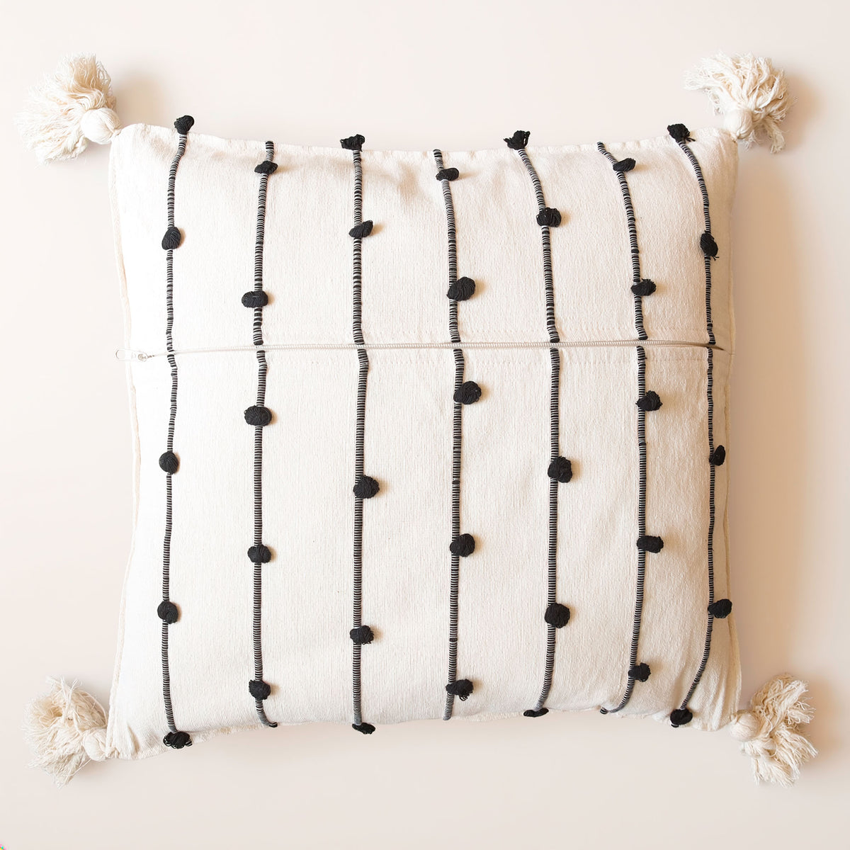 An off-white textured throw pillow with black vertical stripes decorated with pom-poms and tassels, shown against a solid cream background.