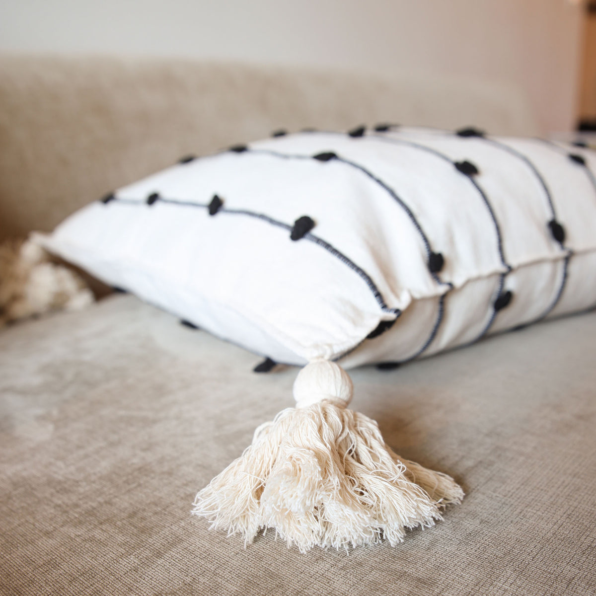 A white throw pillow with black embroidered lines and black pom-pom accents, resting on a beige textured sofa. A large cream-colored tassel is in the foreground.