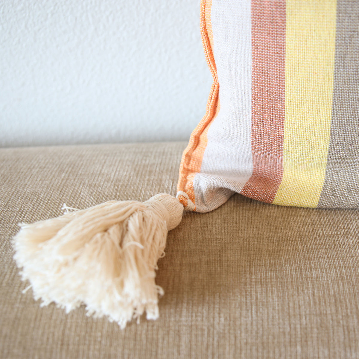 A close-up of a woven throw pillow with vertical stripes in cream, orange, dusty pink, yellow, and gray, featuring a cream tassel on a beige textured sofa.