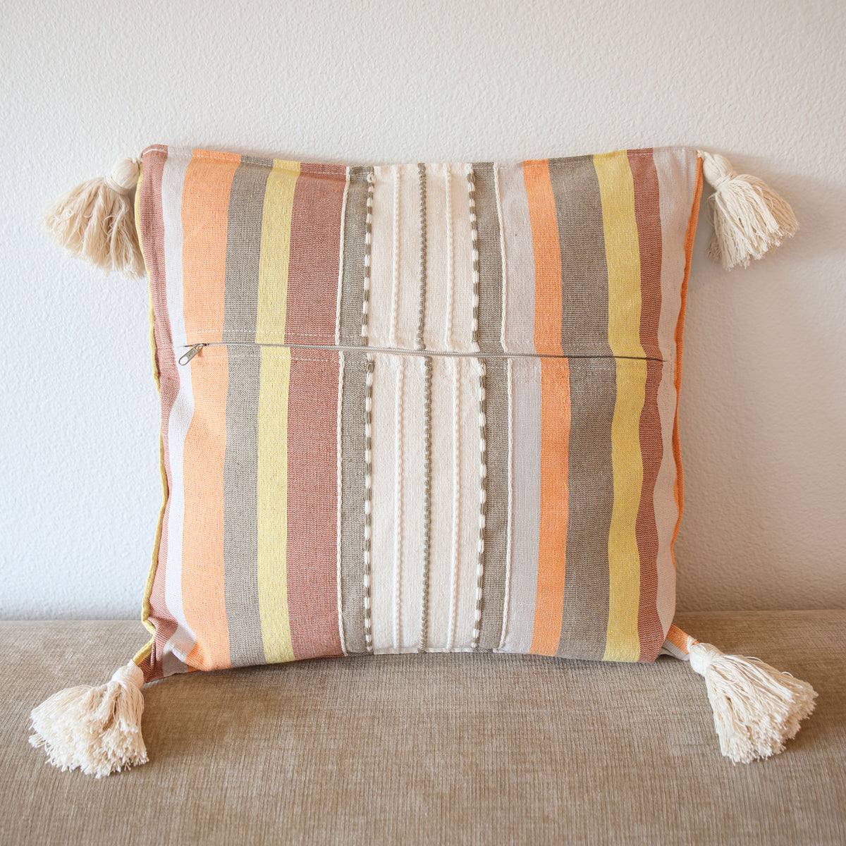 A square decorative throw pillow with vertical stripes in beige, orange, yellow, and brown, featuring cream tassels on two sides and a zippered pocket on the front, set against a neutral background.
