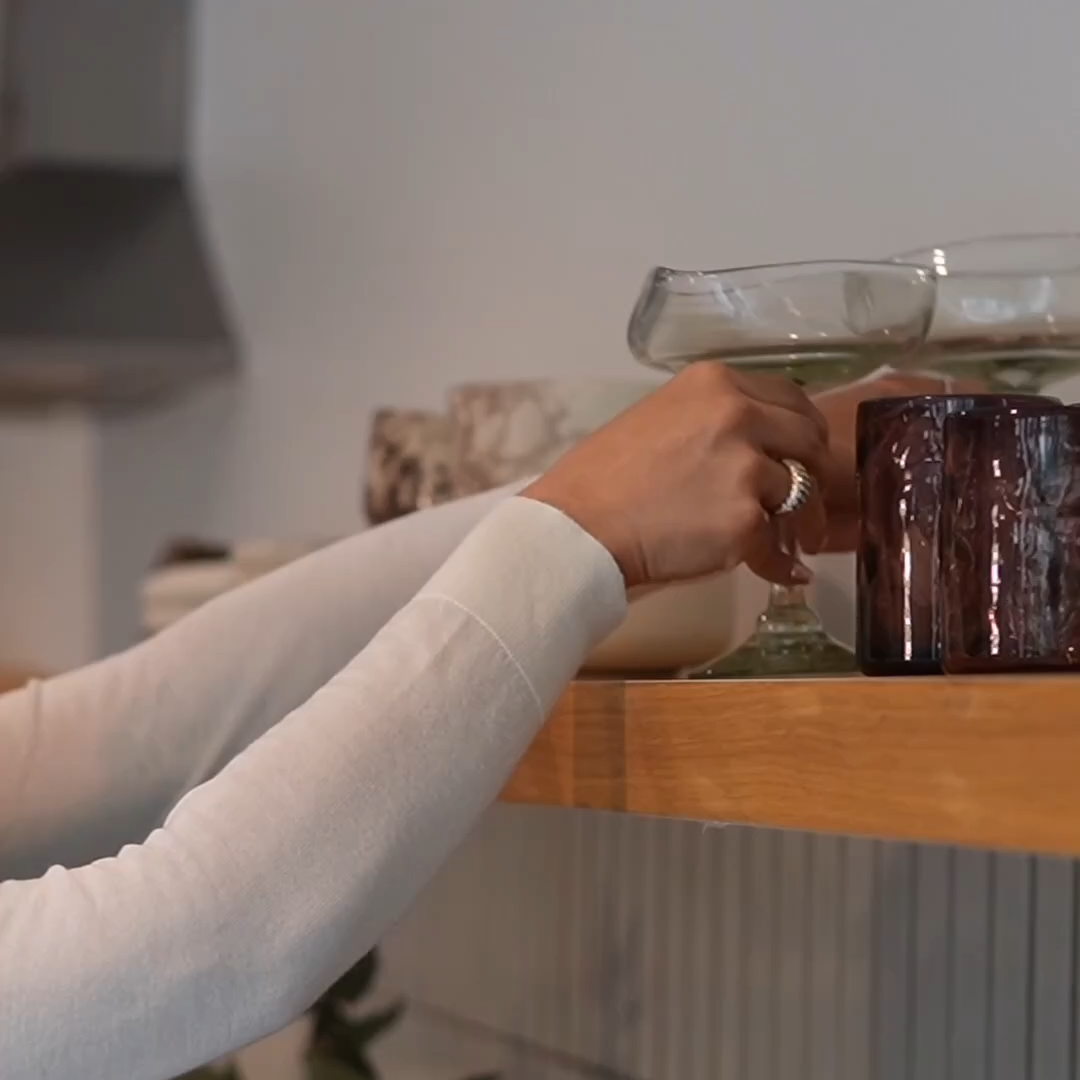 A person wearing a white long-sleeve top reaches for a green glass on a wooden shelf in a modern kitchen setting.