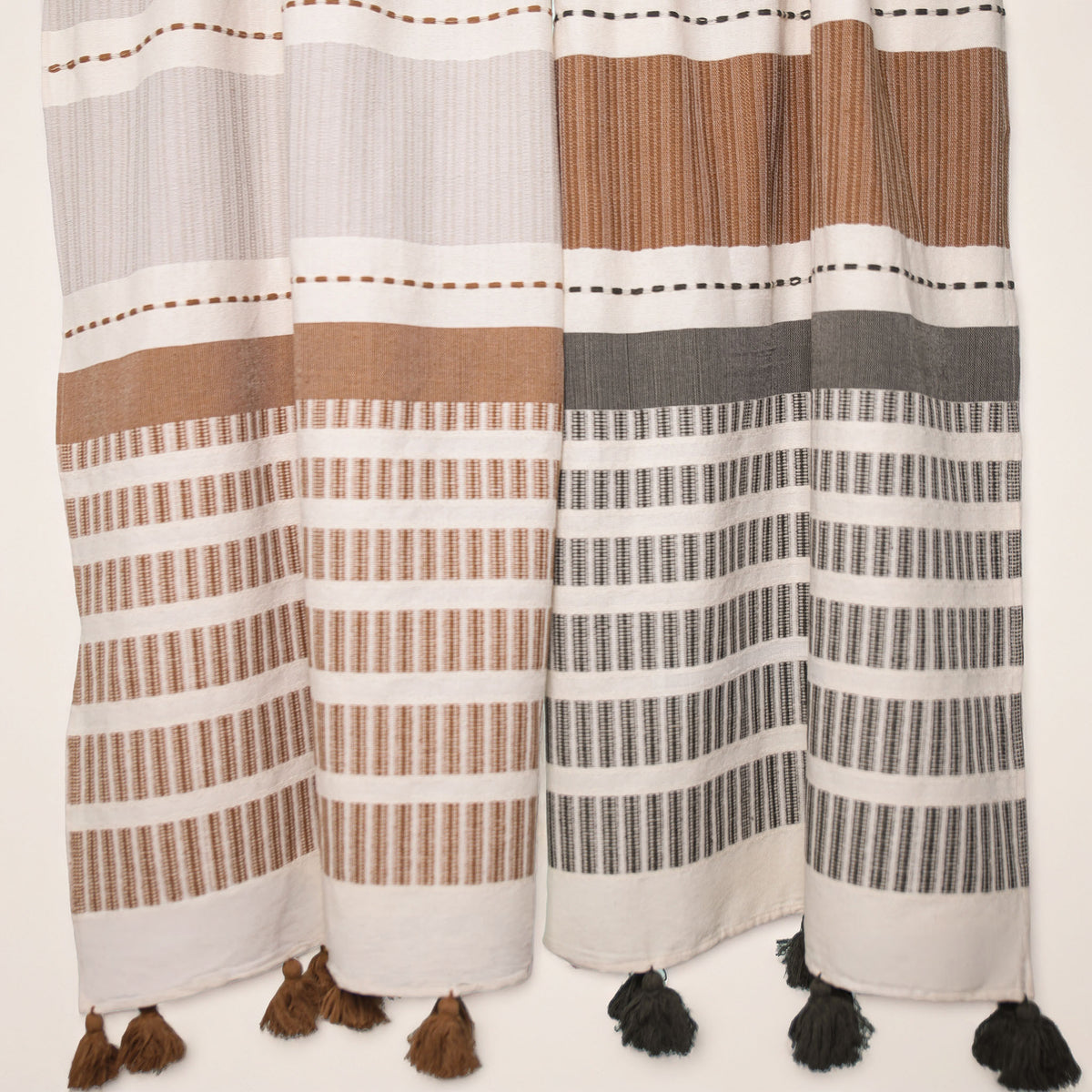Two woven textile throws with horizontal stripes in white, tan, brown, and dark gray, finished with tassels along the bottom edge.