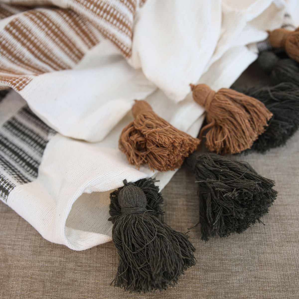 A close-up of a woven textile with fringed tassels, shown draped on a neutral fabric surface. The product appears to be a throw blanket or scarf in off-white, cream, tan, and black woven stripes, with several tassels in tan and black visible.