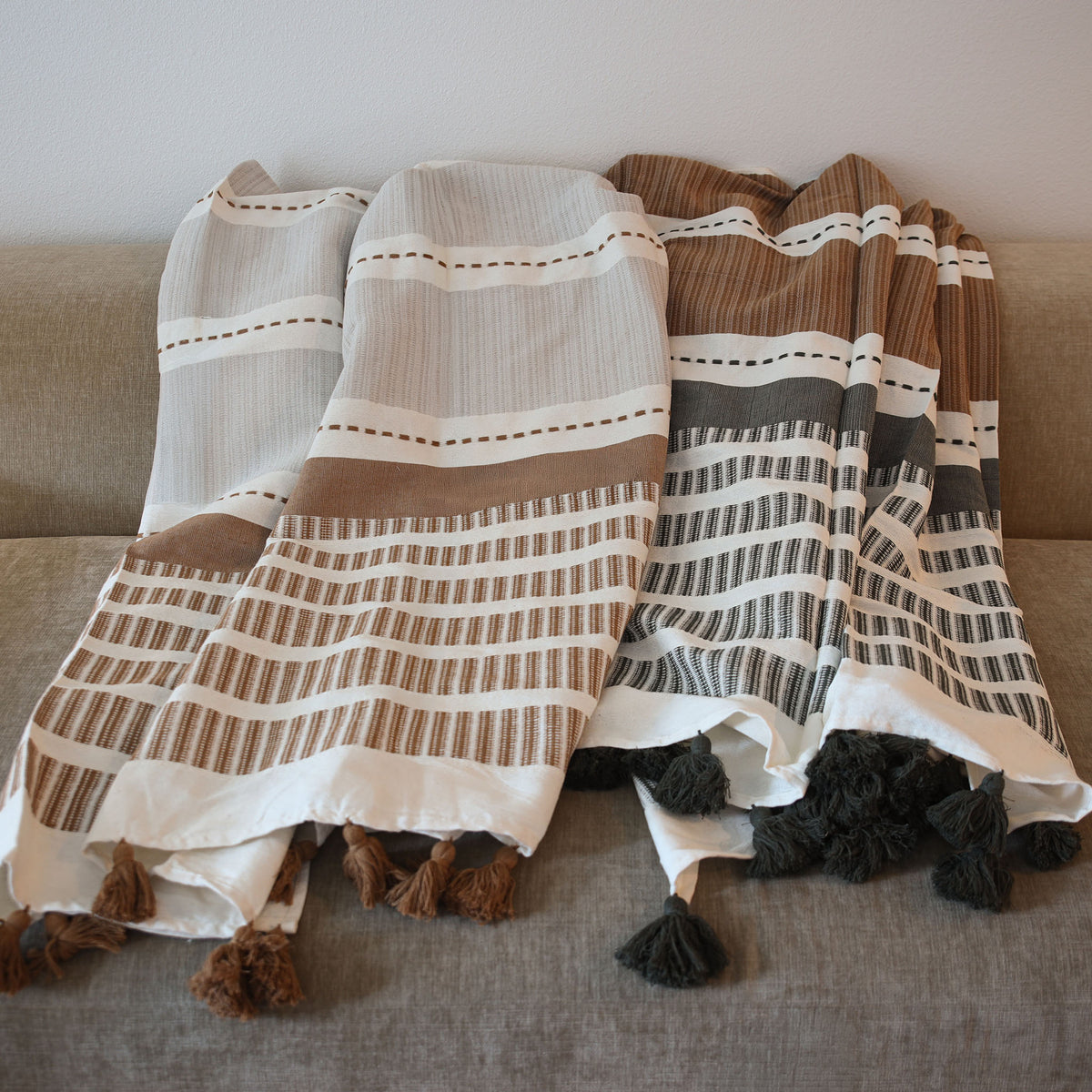 Two woven throw blankets draped over a tan fabric sofa. The blanket on the left is off-white and brown with horizontal stripes and dotted lines; the right blanket is cream, brown, and dark gray with vertical stripes and dark gray tassels.