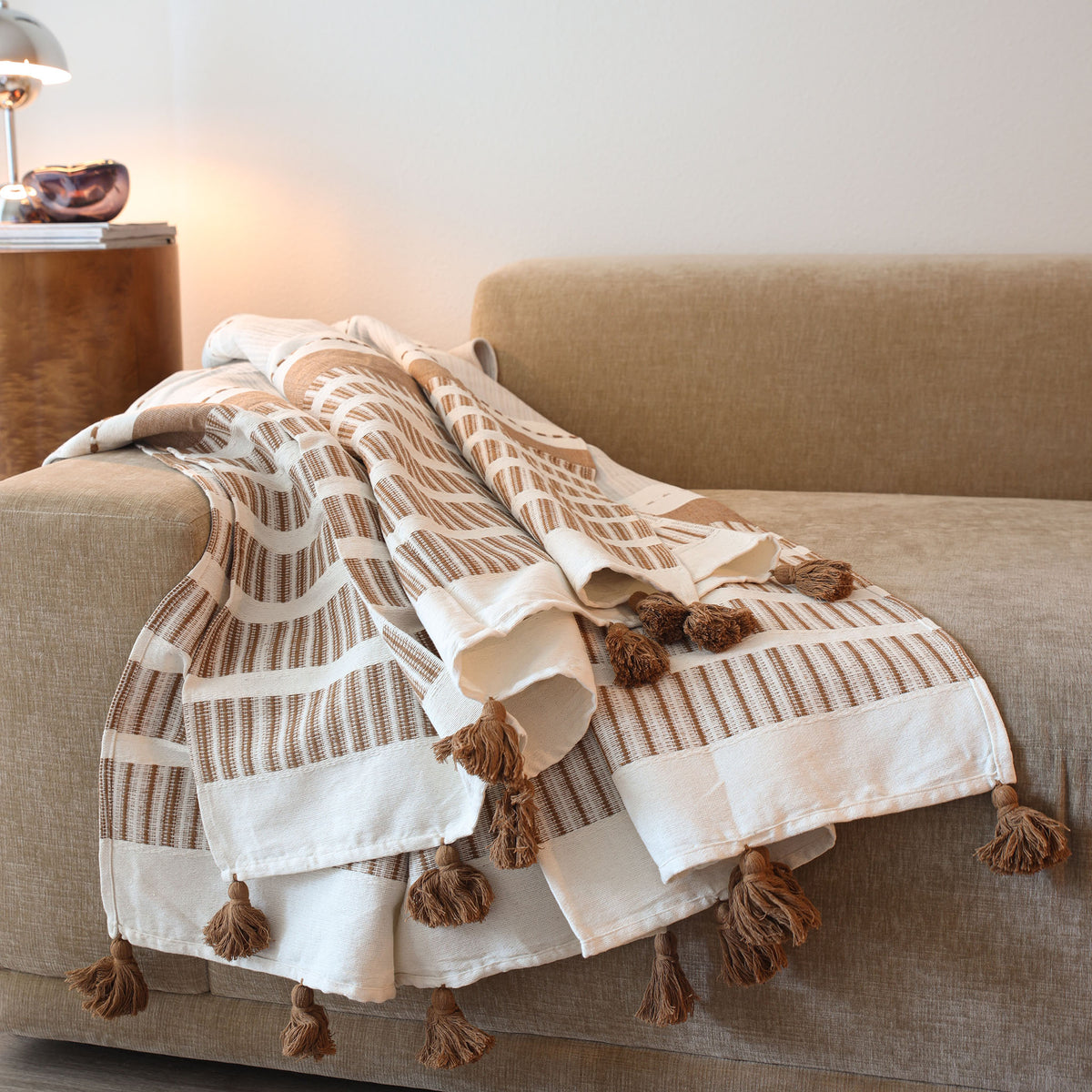 A woven throw blanket with brown and cream stripes and tassels, draped over a beige fabric sofa in a warm, modern living room setting.