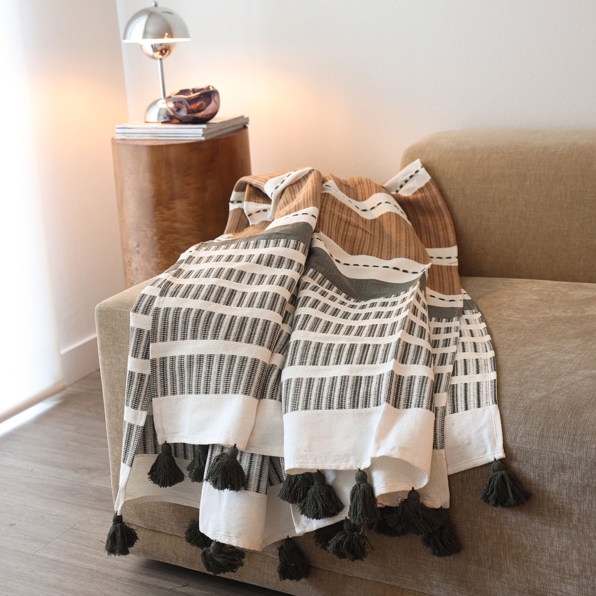 A woven throw blanket with black and white geometric stripes and dark tassels, draped over a beige armchair in a softly lit living room next to a wooden side table.