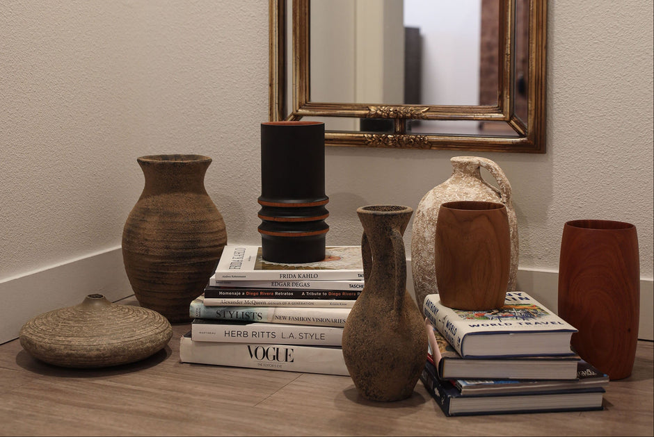 A styled collection of rustic ceramic and wooden vases arranged on a wooden floor in front of a gold-framed mirror, with books used as styling props.
