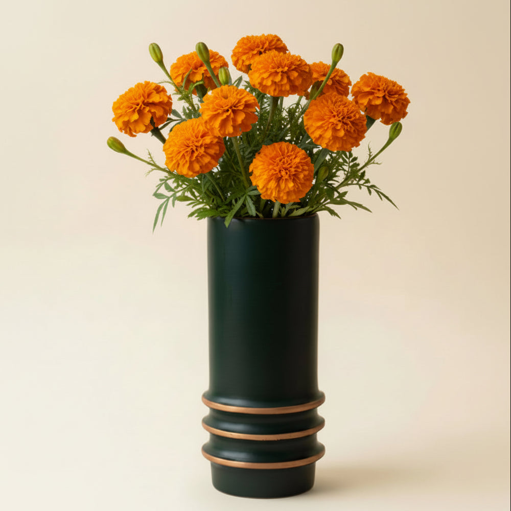 A black cylindrical wood planter with stacked tan rings near the base, holding a orange marigold floral arrangement, set against a beige wall.